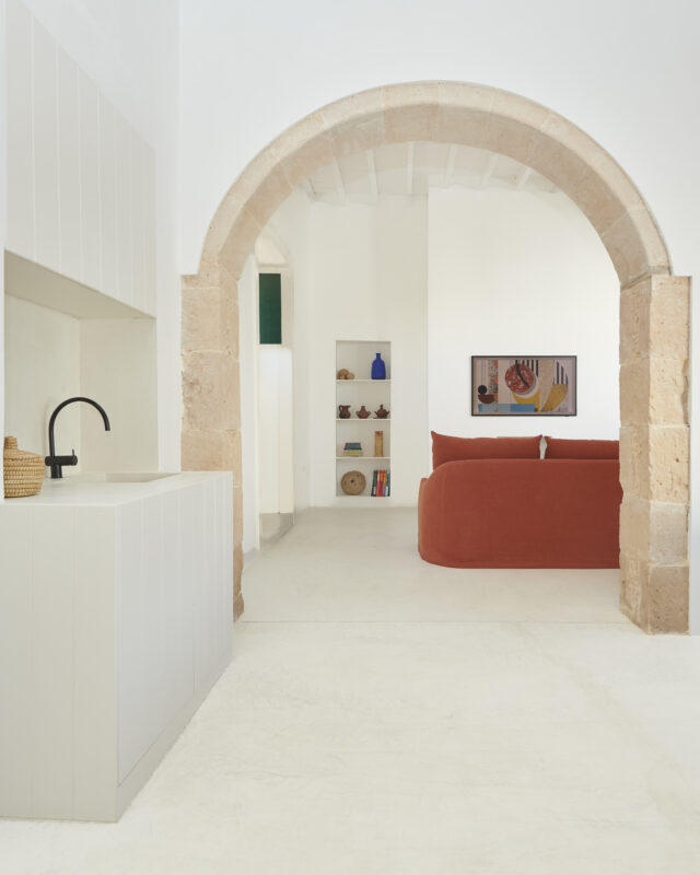 Bright, minimalist interior of Casa Montelongo featuring a curved stone arch, a white kitchen, and a red sofa.