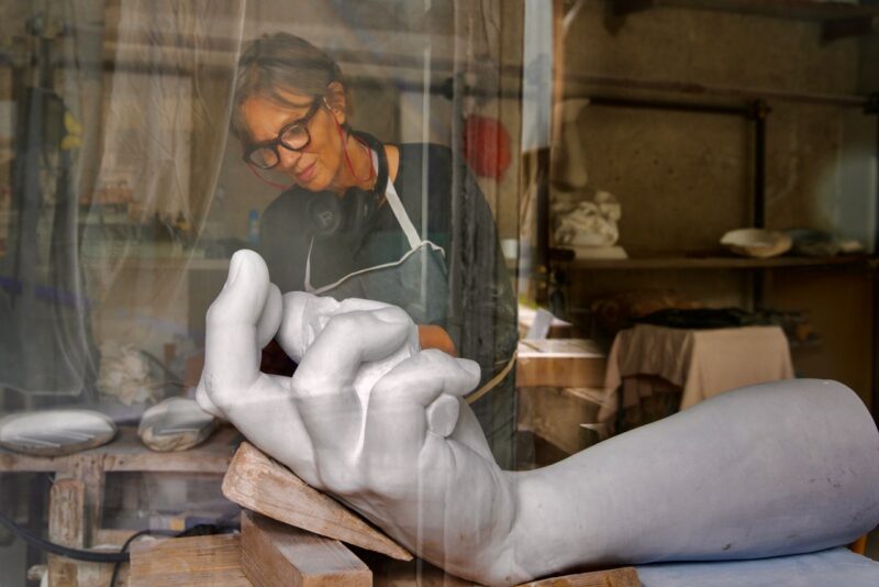 Artist working on a large white sculpture of a hand in a studio, with tools and materials visible in the background.
