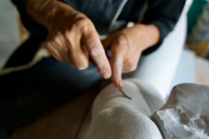Hands carving a sculpture, focusing on a tool against a white surface, with a blurred background.