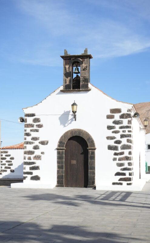 Small white chapel with a bell tower and stone accents, set against a blue sky in La Oliva, Fuerteventura.