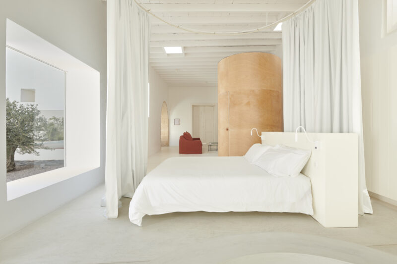 Minimalist bedroom with a white bed, draped curtains, and a round orange structure, featuring a view of nature outside.