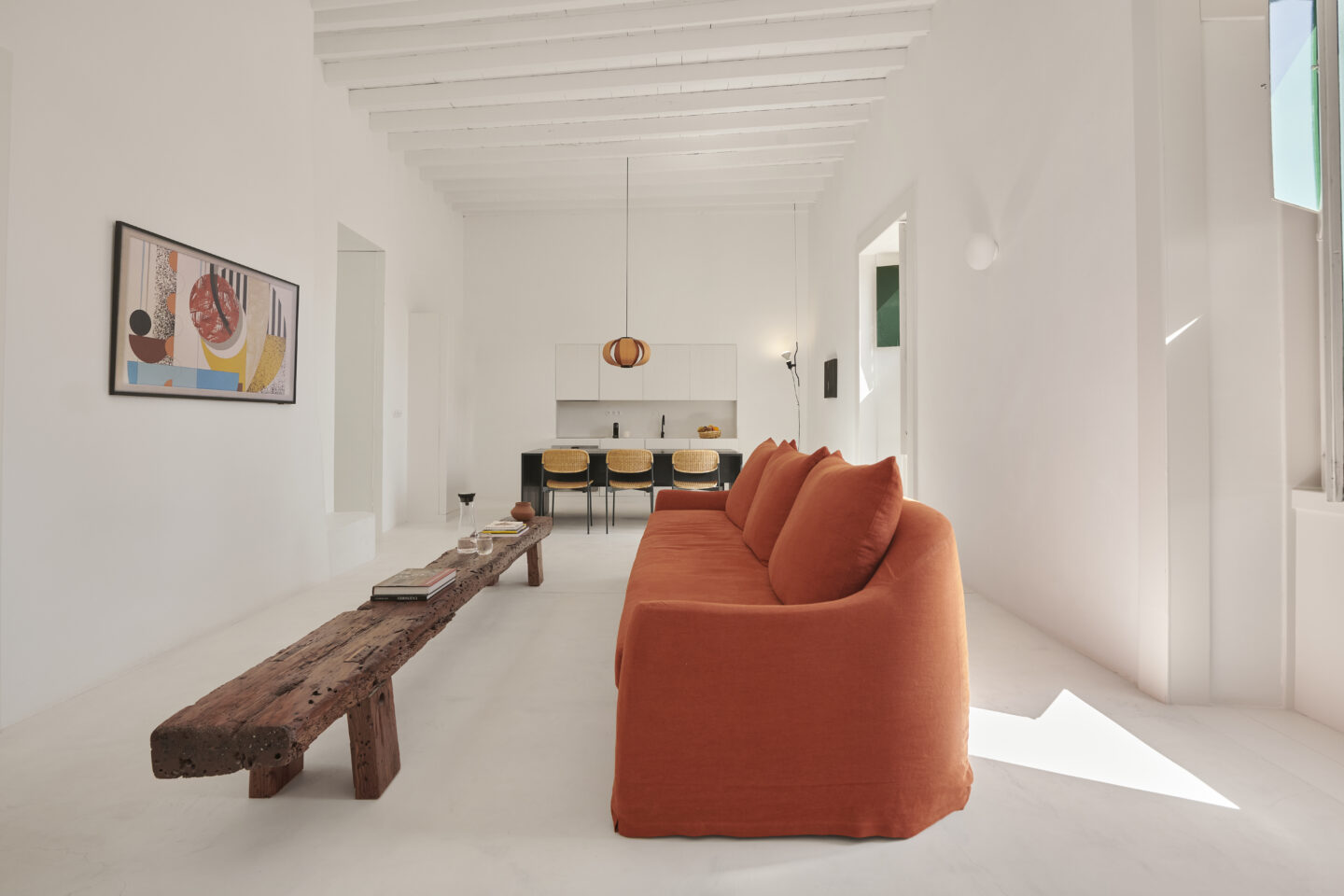 Bright living room with white walls, an orange sofa, a wooden coffee table, and a minimalist kitchen in the background.