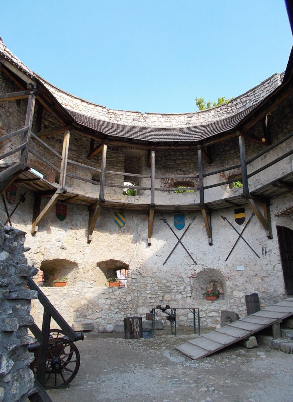 Interior view of a stone structure with a circular layout, featuring wooden beams, shields, and a stone floor.