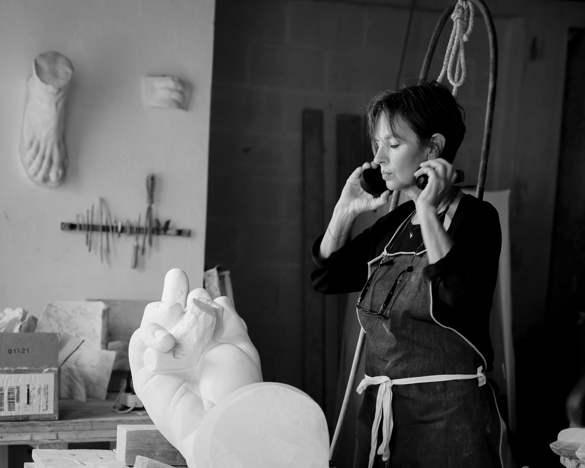 Artist in an apron using a phone, surrounded by sculpting tools and a large white hand sculpture in a studio.