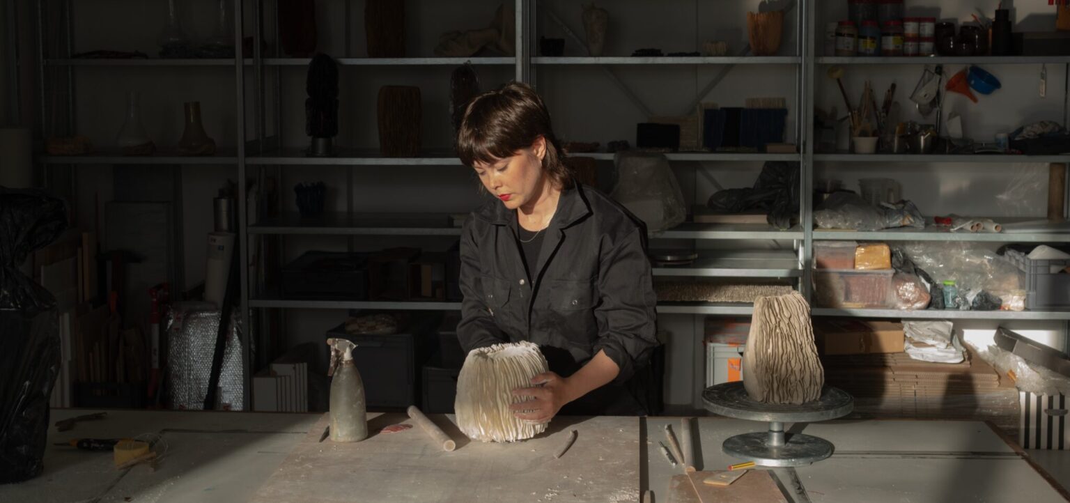 Juliana Maurer working in her studio, shaping a ceramic piece on a table with shelves of materials in the background.