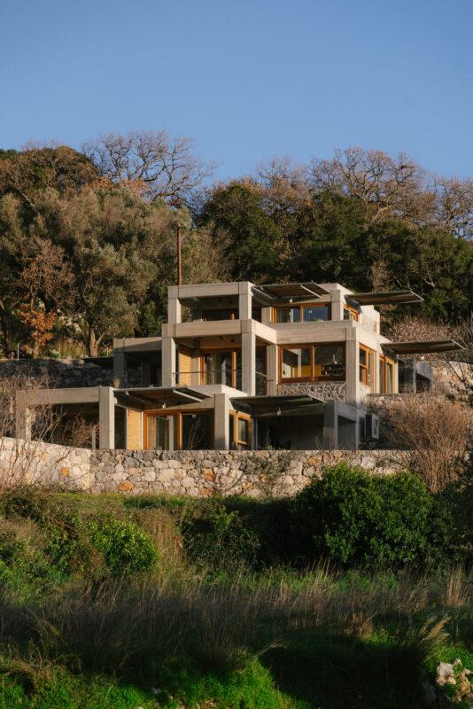 Modern multi-level house with large windows, surrounded by trees and greenery, set against a clear blue sky.