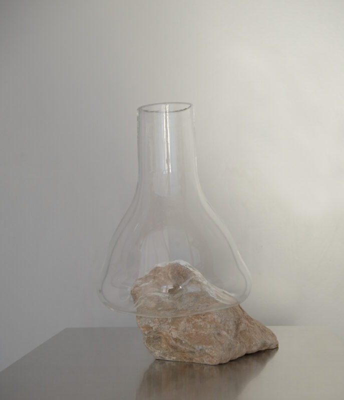 Glass laboratory flask resting on a rock, set against a light-colored wall.