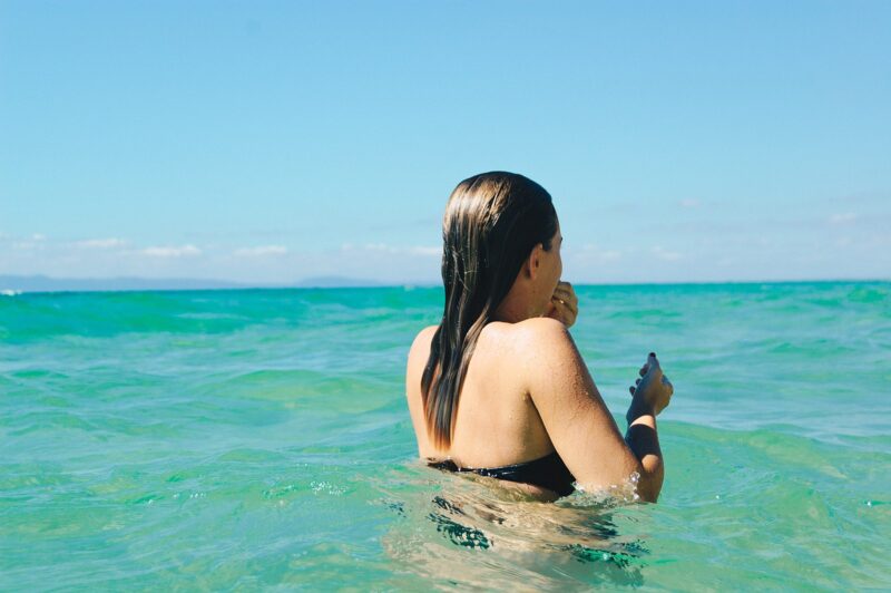 Woman in a black bikini wading in clear turquoise water under a bright blue sky in Melides, Portugal.