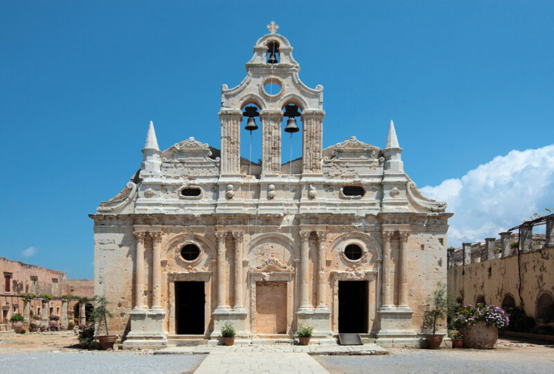 Facade of Moni Arkadiou, a historic monument in Crete, featuring ornate architecture and bell towers against a blue sky.