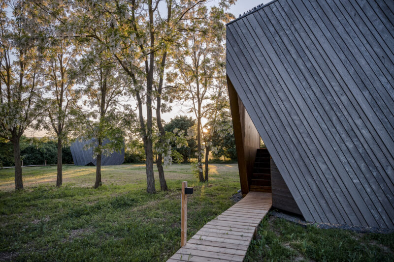 Modern cabin with angled wooden exterior, surrounded by trees and a pathway leading to the entrance, at sunset.