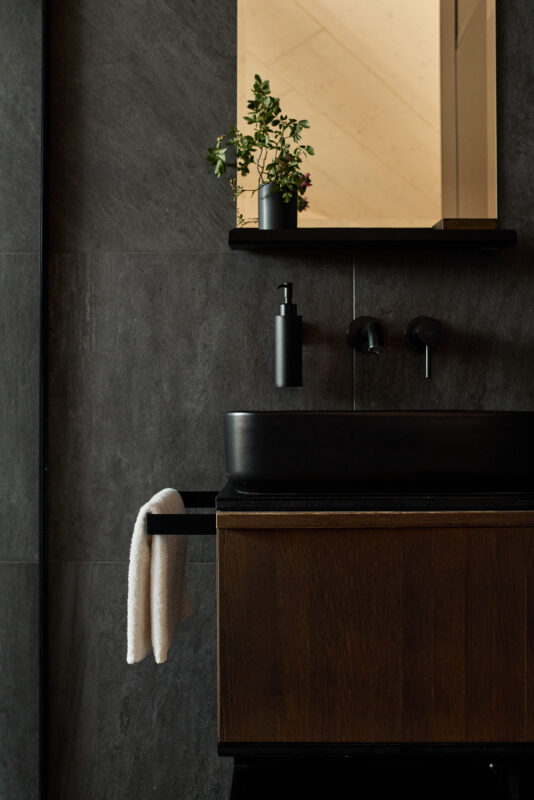 Modern bathroom featuring a dark stone wall, a sleek black sink, and a wooden cabinet, with a small plant on a shelf.