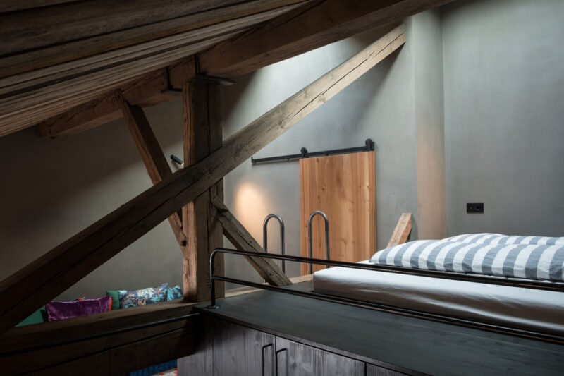 Interior view of a loft with wooden beams, a bed with striped bedding, and a sliding door in a modern design.