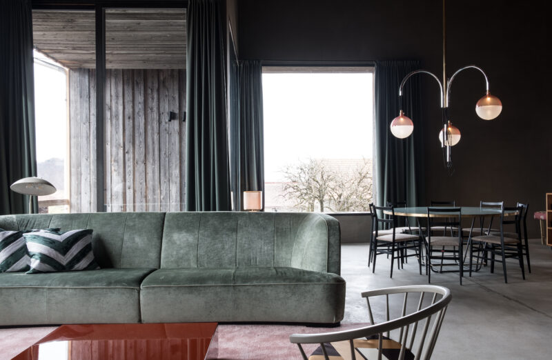 Modern interior of a barn with a green velvet sofa, dining area, and large windows showcasing a cloudy landscape.