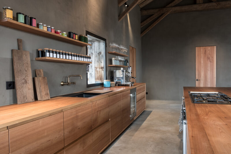 Modern kitchen with wooden cabinetry, open shelving displaying jars, and a minimalist design in a rustic setting.
