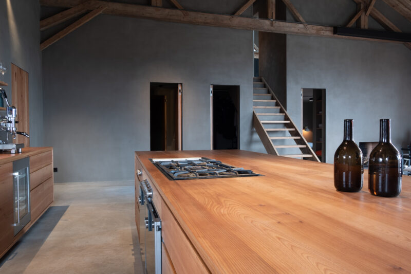Modern kitchen with wooden island, gas stove, and dark glass bottles; minimalist design in a spacious, industrial-style room.