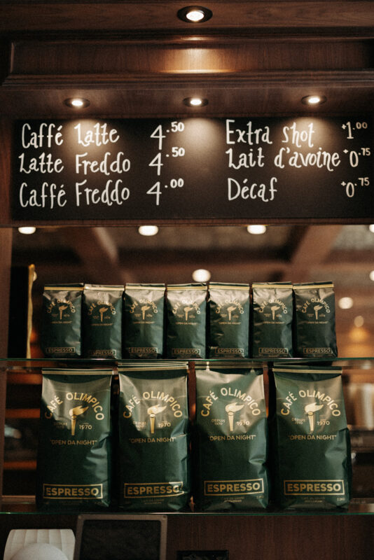 Menu board listing coffee prices above bags of Cafe Olimpico espresso on a wooden counter.