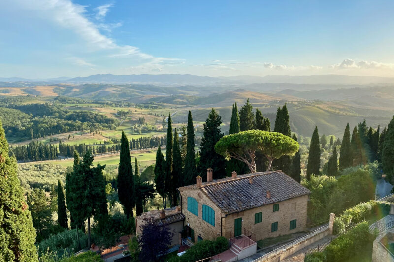 Tuscan landscape featuring rolling hills, cypress trees, and a rustic stone house under a clear blue sky.