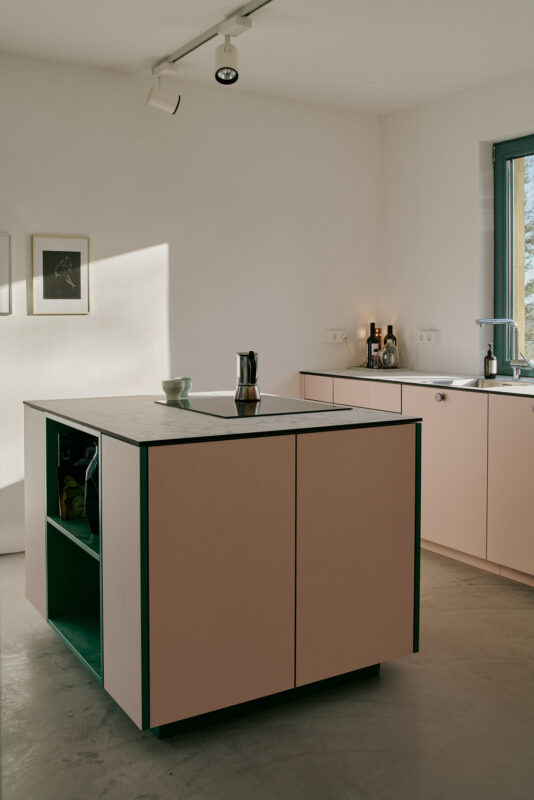 Modern kitchen featuring a pink island with green accents, minimalist design, and natural light streaming through a window.