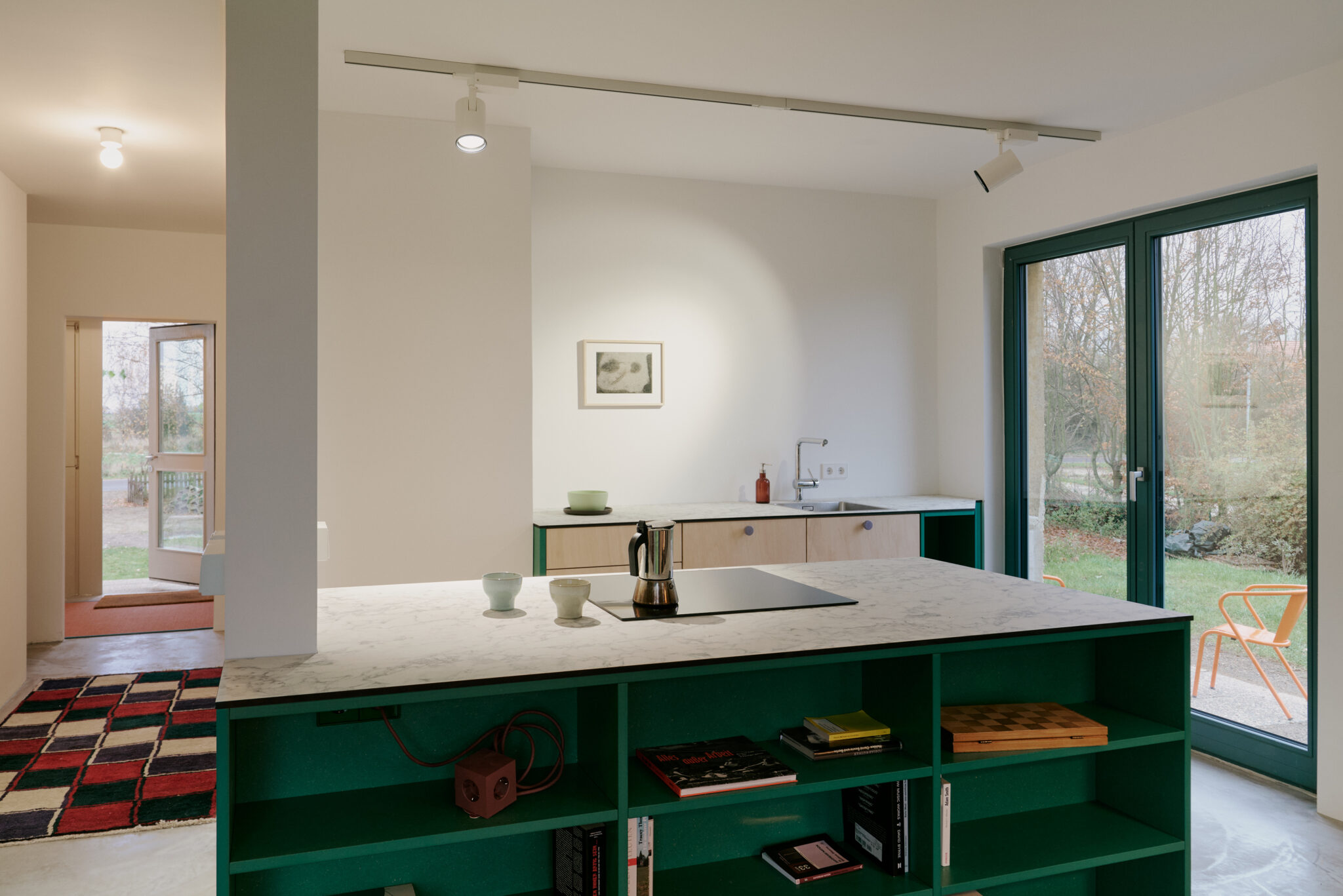 Modern kitchen with green shelving, marble countertop, and a view of a garden through large glass doors.