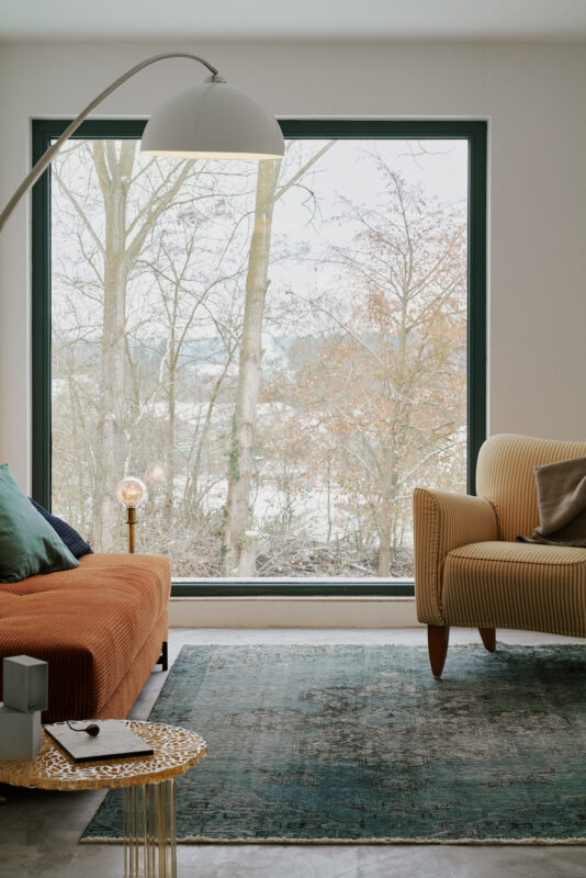 Cozy living room with an orange sofa, striped armchair, and large window revealing a view of trees and a cloudy sky.