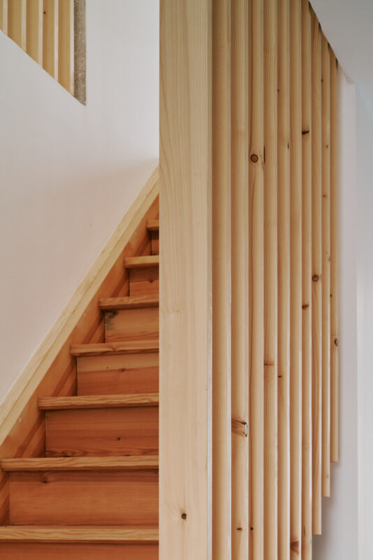 Wooden staircase with light-colored wooden steps, flanked by vertical wooden slats against a white wall.