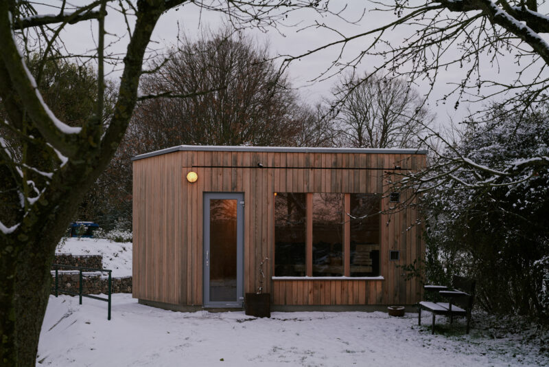 Modern wooden cabin with large windows, surrounded by snow-covered ground and trees, at dusk.