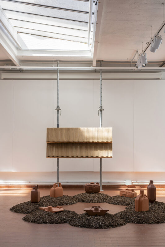Art installation featuring a wooden structure suspended above a circular arrangement of clay vessels on a gravel bed.