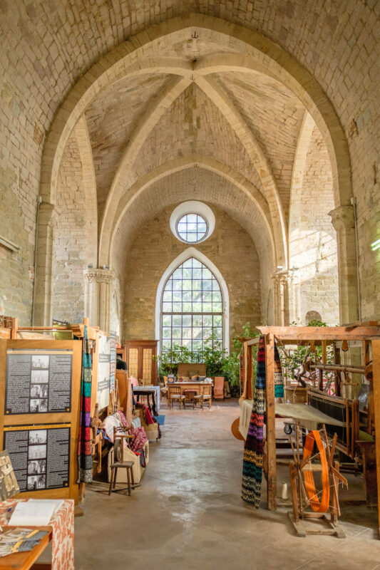 Interior of a stone building with high arched ceilings, featuring tables, textiles, and plants.
