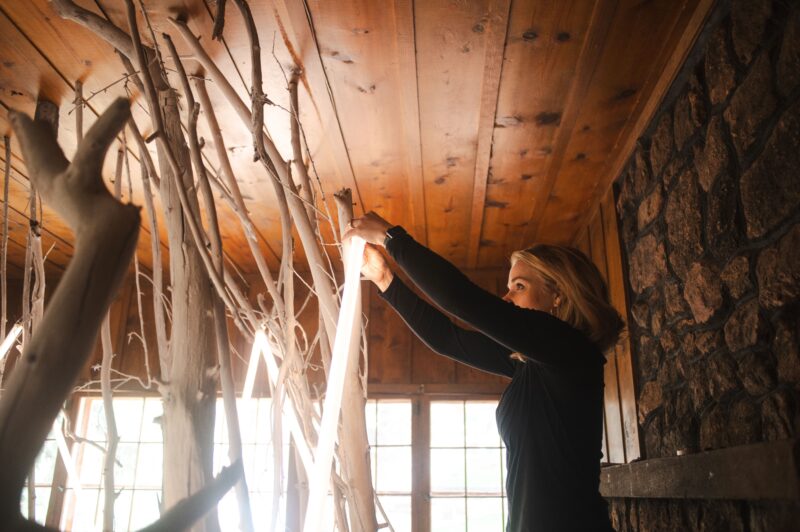 Woman arranging branches in a wooden room with stone walls, sunlight filtering through windows.