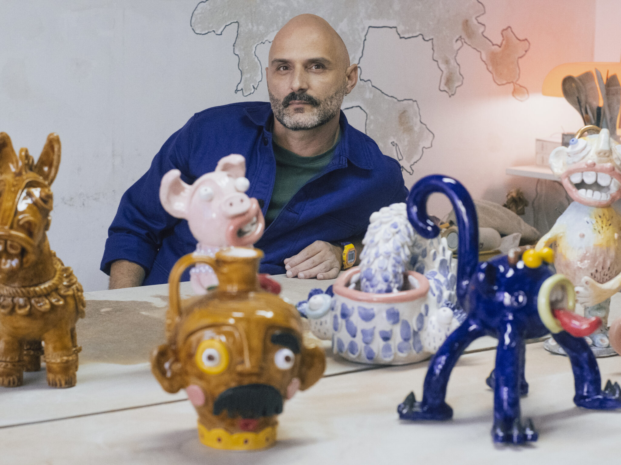 Carlos Manuel Gonçalves sits at a table surrounded by colorful ceramic sculptures in his atelier.
