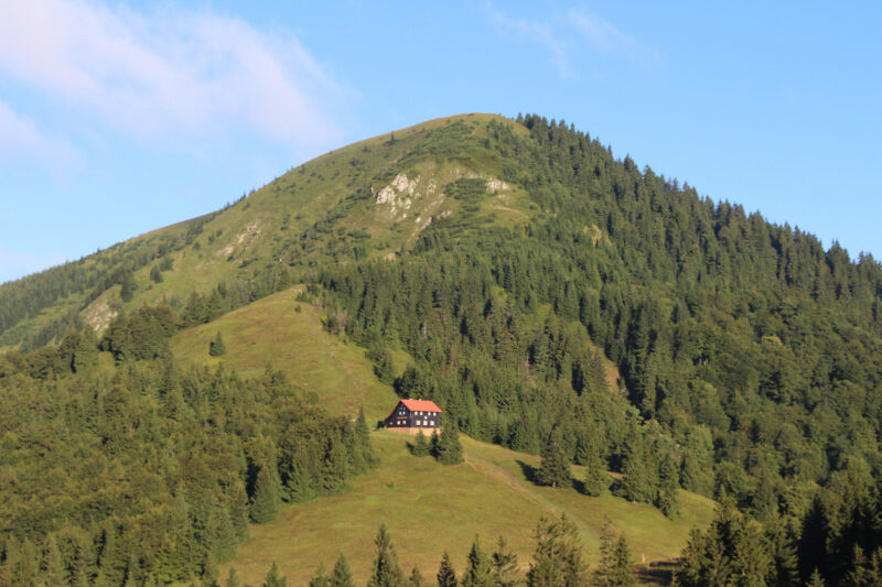 Lone wooden cabin with a red roof surrounded by lush green hills and forested mountains in Velká Fatra, Slovakia.