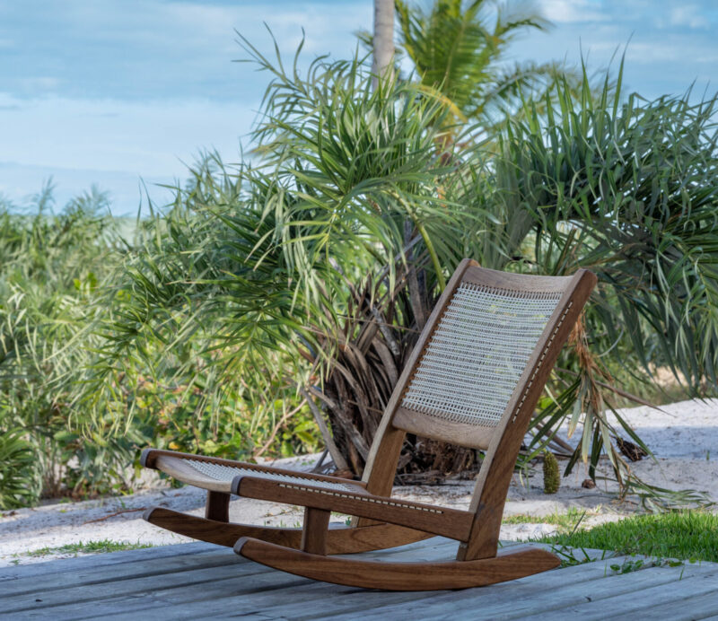 Wooden rocking chair on a deck surrounded by tropical plants and a view of the ocean in the background.