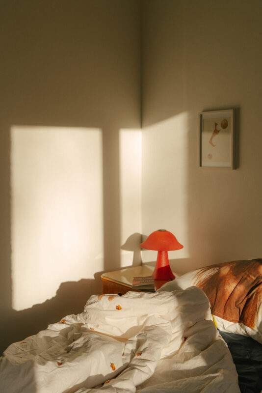 Sunlight casts shadows on a bed with rumpled white sheets, a red lamp, and a framed artwork on the wall.