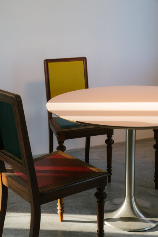 Round pink table with a metallic base, flanked by two wooden chairs with colorful upholstery in green and red.