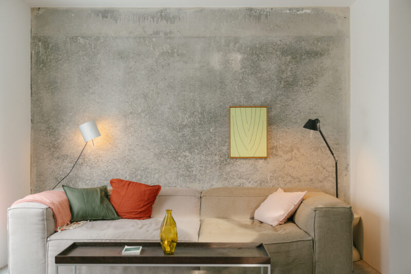 Modern living room with a beige sofa, colorful cushions, a glass vase, and abstract wall art against a textured gray wall.