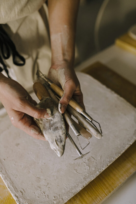 Hands holding sculpting tools and a clay piece over a work surface.
