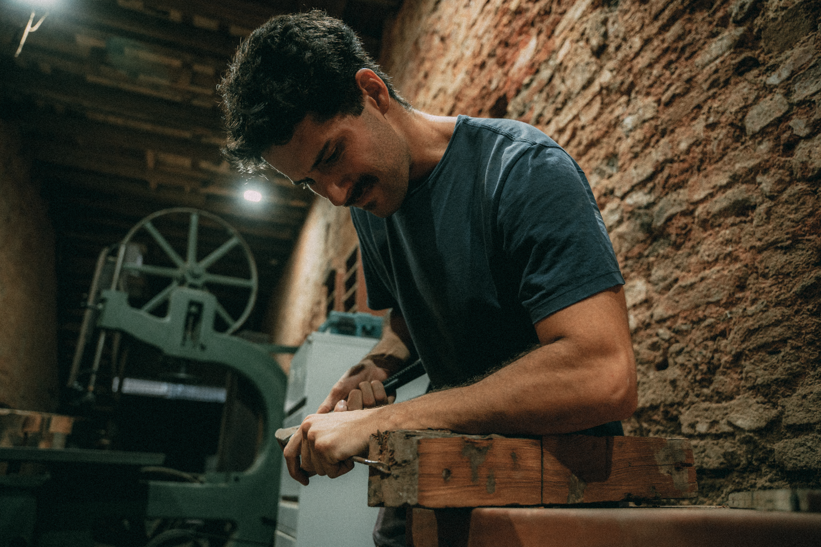 Man working with wood in a workshop, focused on carving a piece of wood with tools.