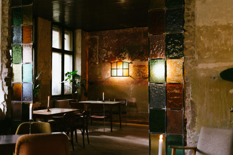 Dimly lit restaurant interior with colorful glass paneling, wooden tables, and candles on the tables.