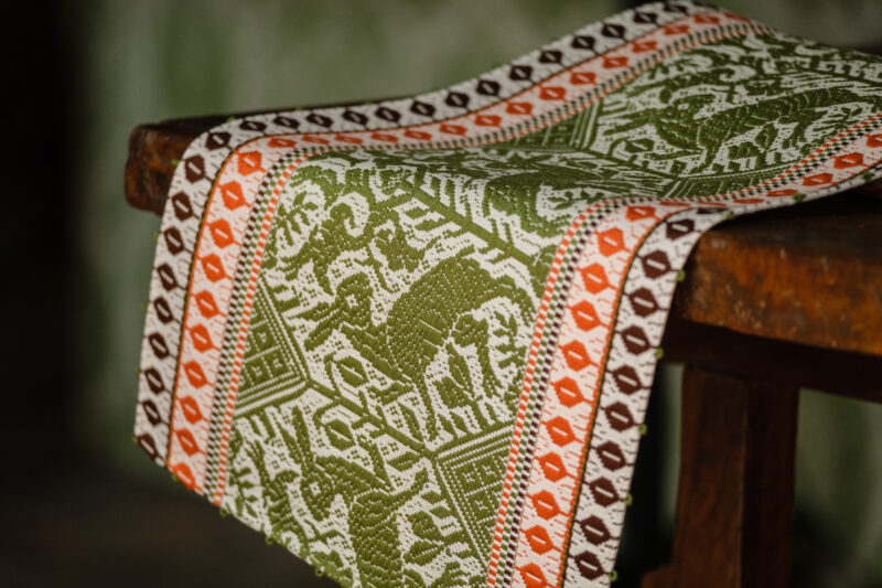 Colorful woven textile featuring intricate patterns in green, orange, and brown, draped over a wooden stool.