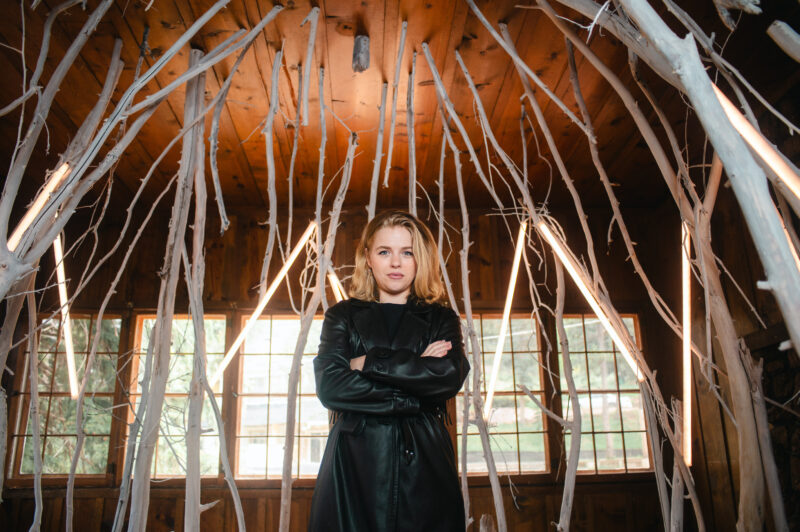 Woman in a black leather coat stands confidently among white branches in a wooden room with large windows.