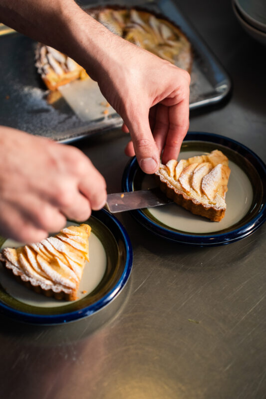 Slicing a dessert tart with apple slices on two plates in a kitchen setting.