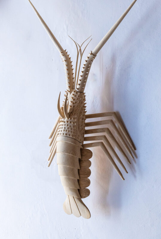 Wooden sculpture of a lobster mounted on a white wall, showcasing intricate details and textures.