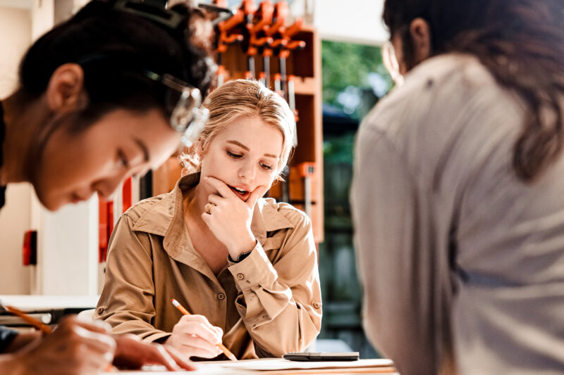Three individuals engaged in a creative discussion, with one woman thoughtfully observing while others write notes.