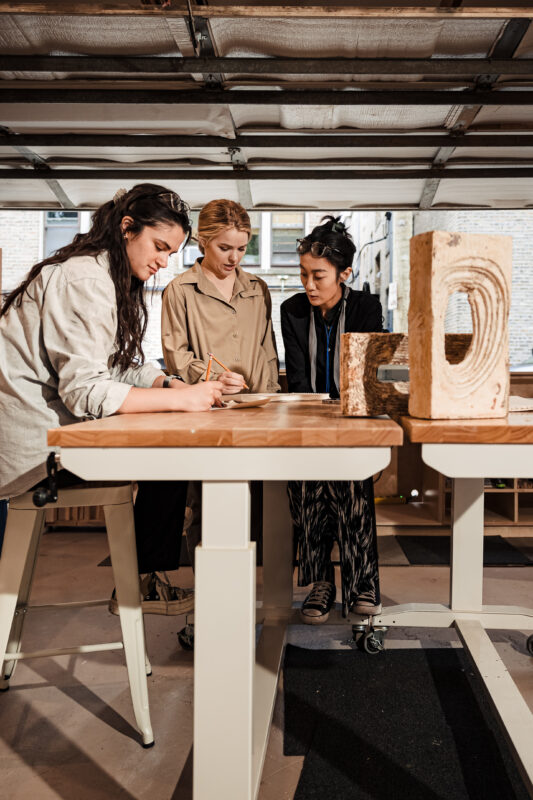 Three women collaborate at a workspace, sketching and discussing designs, with wooden blocks nearby.