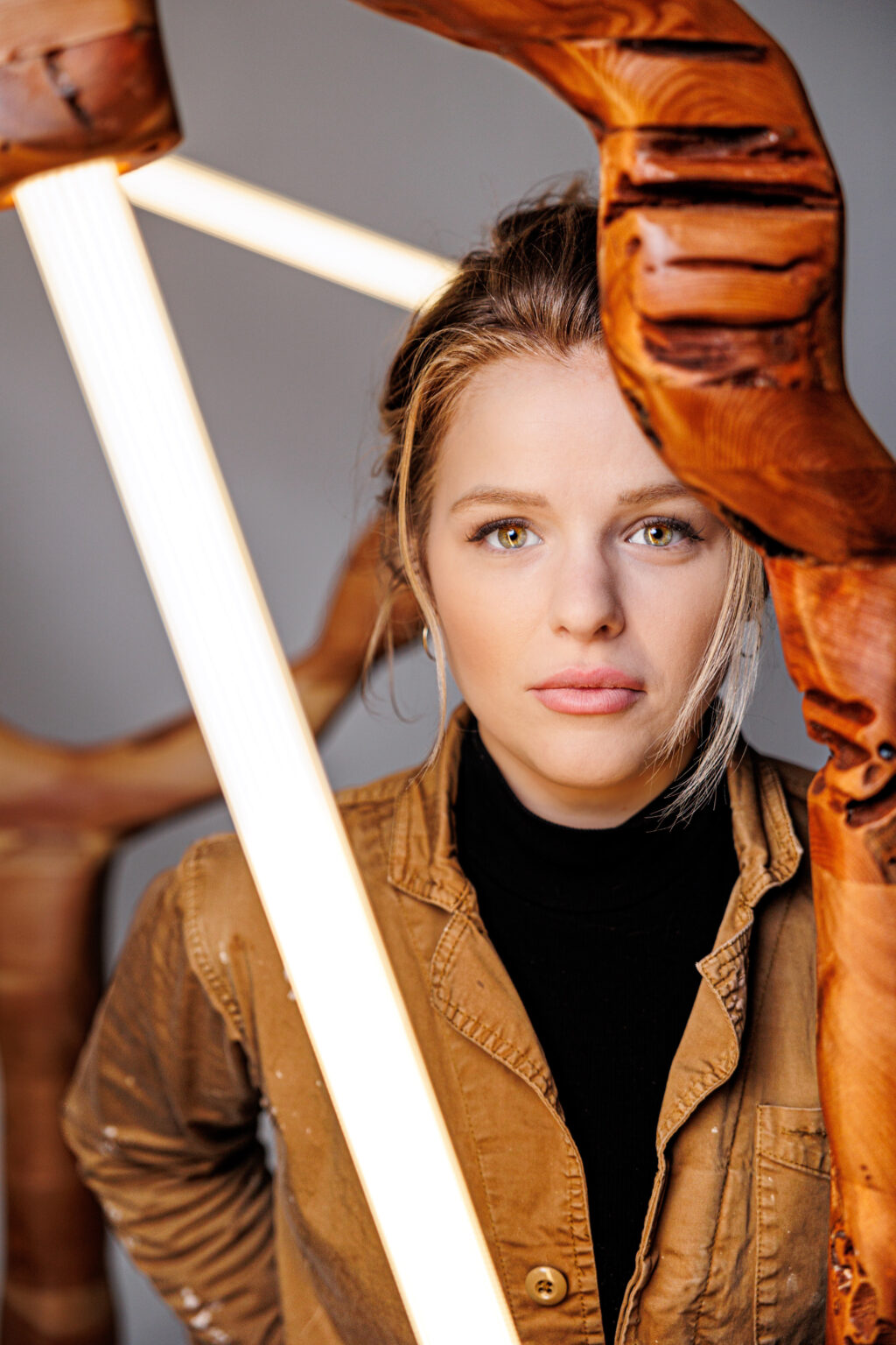 Woman with blonde hair and a serious expression, framed by a wooden sculpture and illuminated by a light.