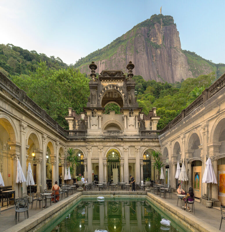 Historic architecture surrounds a tranquil pool, with a mountain and statue in the background, in Rio de Janeiro.