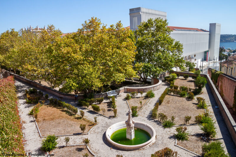 Garden scene in Lisbon, Portugal, featuring a circular fountain surrounded by trees and shrubs under a clear blue sky.