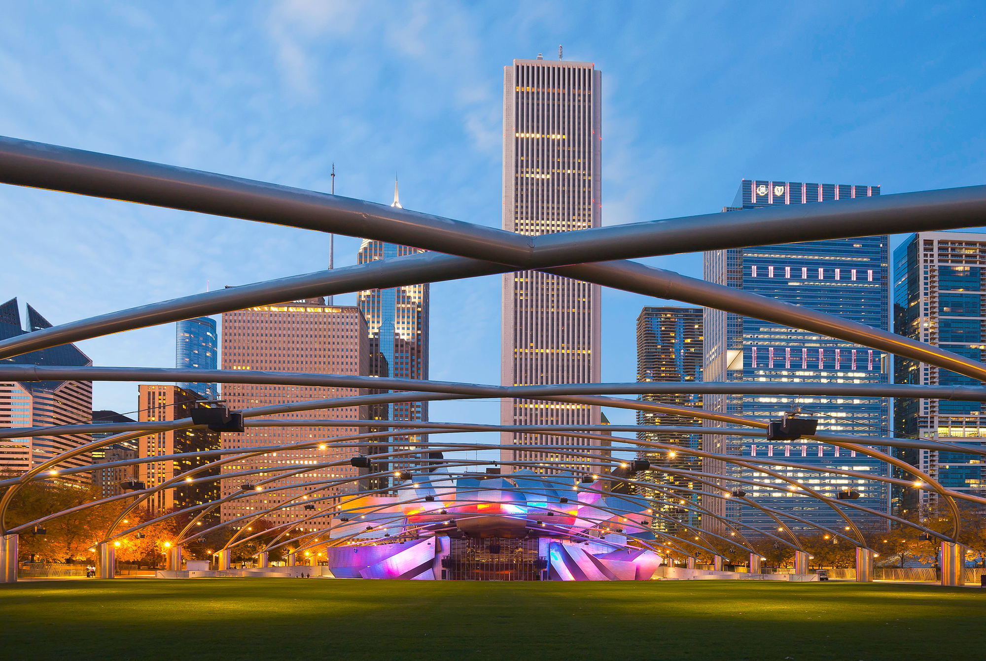 Jay Pritzker Pavilion in Chicago, illuminated at dusk, with city skyscrapers in the background.
