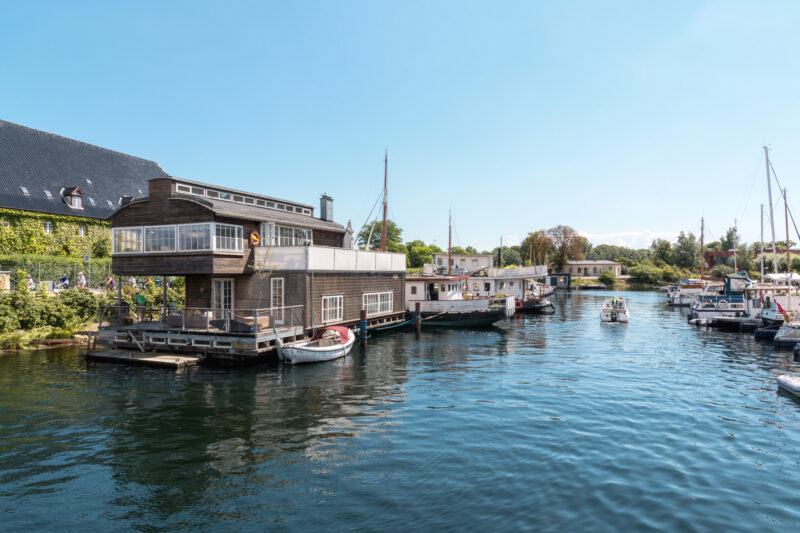 Wooden house on the water in Christianshavn, Copenhagen, with boats docked and clear blue skies above.