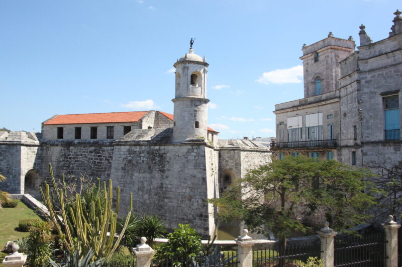 Historic stone building with a tower, surrounded by greenery and blue sky, part of La Giraldilla in Havana.